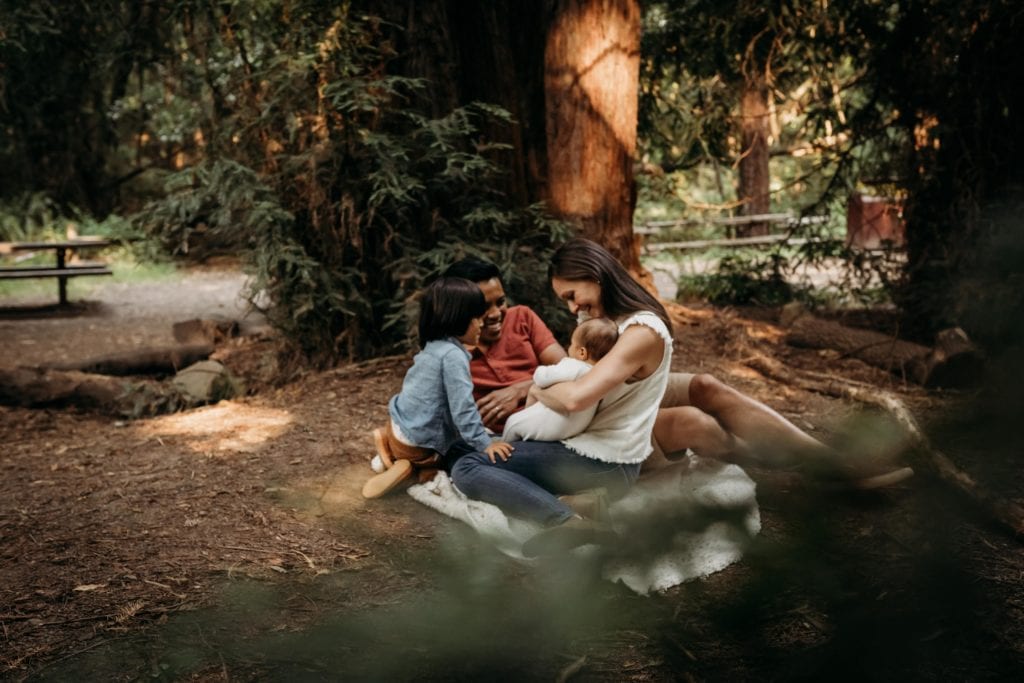 Family of four snuggle on a blanket in grove of trees during family photography session with Xilo Photography at Redwood Regional Park in Oakland, CA.