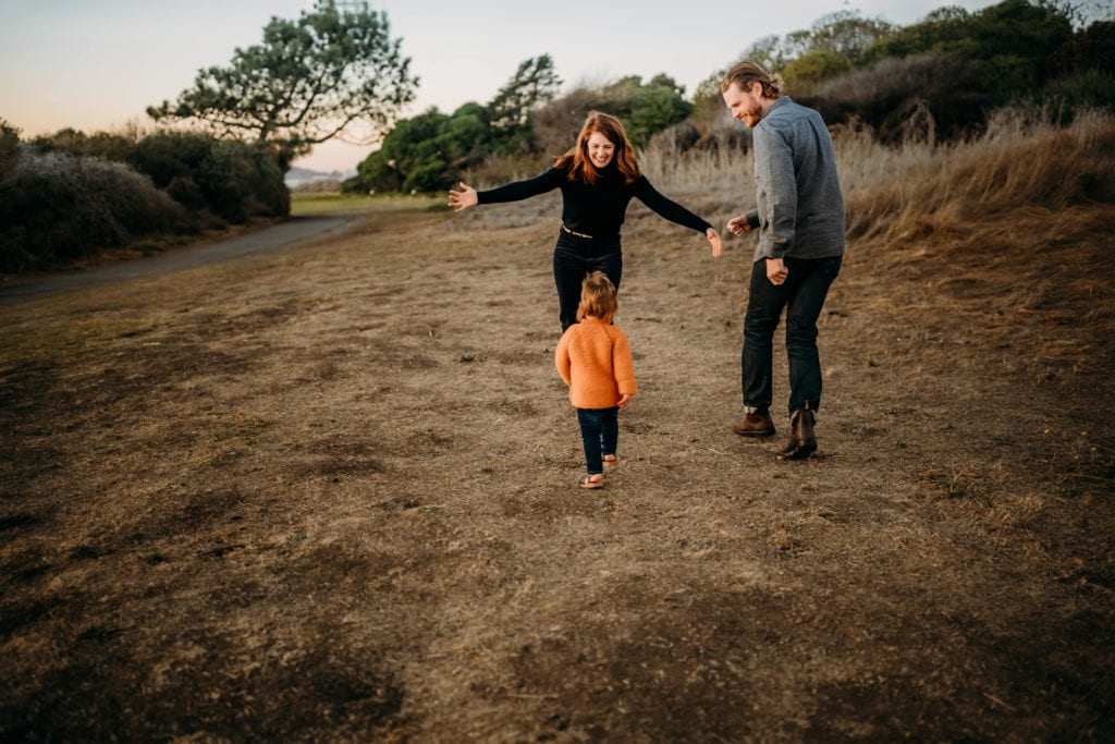 Toddler runs toward laughing parents in an open space at sunset during photography session with Xilo Photography at Berkeley Marina.