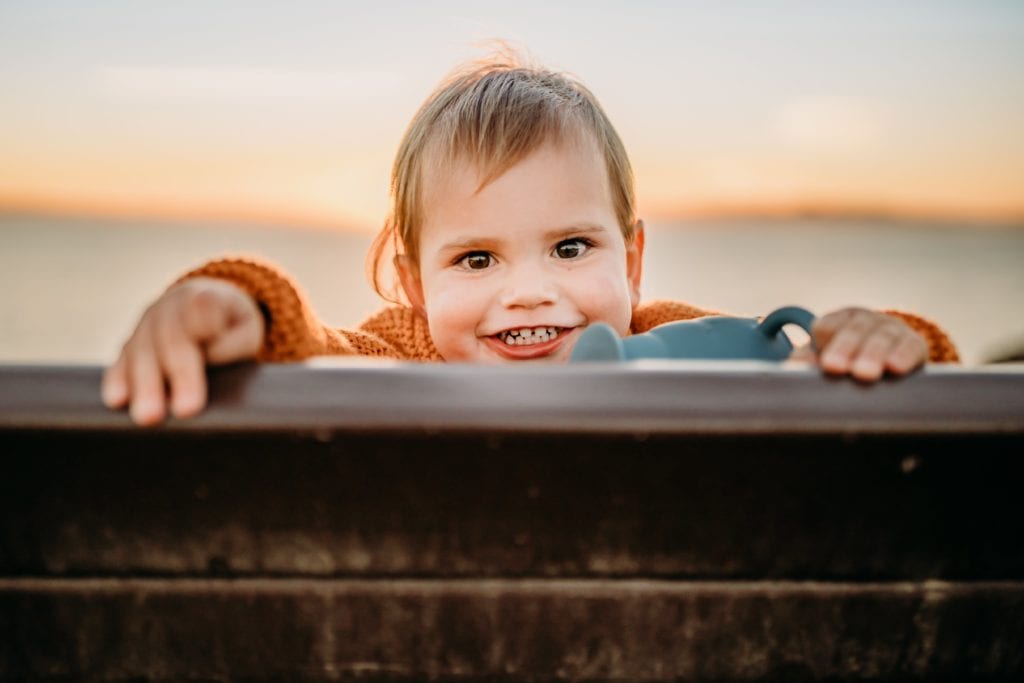 Toddler peaks over bench with sunset in background during a family photography session with xilo photography at Berkeley Marina in Berkeley, CA.