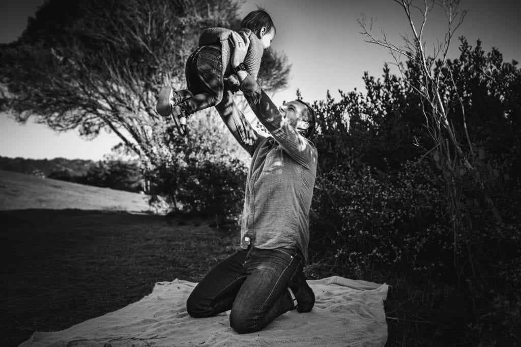 Father holding child up in the air on a blanket in a park during a family photography session iwth Xilo Photography in Berkeley, CA.