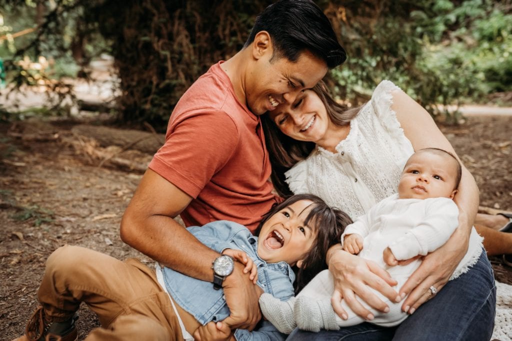 Family of four smile and cuddle together during family photography session with Xilo Photography at Redwood Regional Park in Oakland, CA.