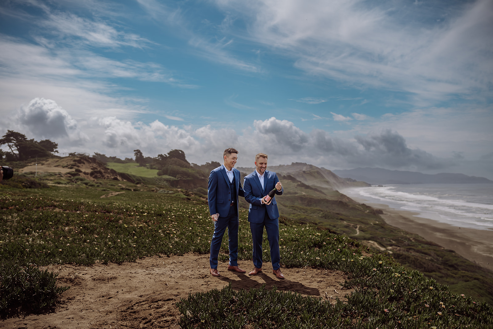 This is a picture of a same sex gay couple on their wedding day/elopement day as the two grooms open a champagne bottle to pour themselves champagne in their glasses while they celebrate their wedding day in their classy blue wedding tuxes at Fort Funston, CA which was their intimate wedding venue. This photo was taken by an inclusive and intimate wedding and elopement photographer, Xilo Photography, Kati Douglas who is based in Oakland, California in the bay area.