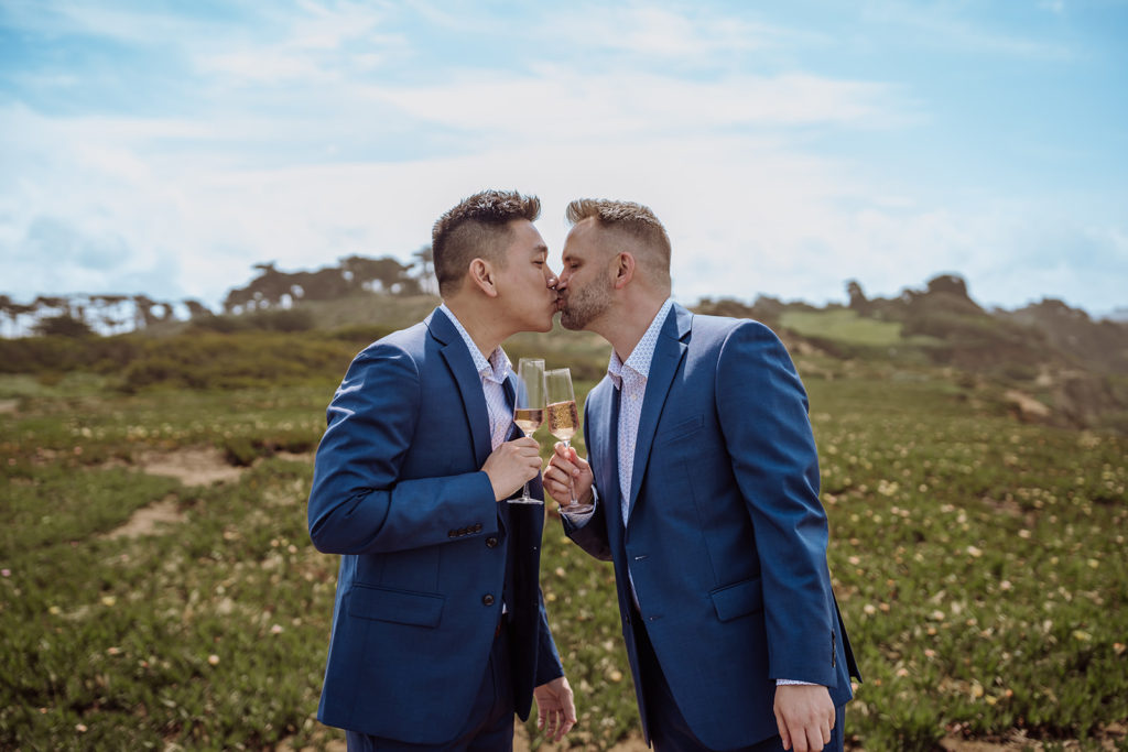 An inclusive and intimate wedding and elopement photographer, Xilo Photography, Kati Douglas who is based in Oakland, California in the bay area, took this picture of a same sex gay couple on their wedding day/elopement day as the two grooms pour themselves champagne so they celebrate their wedding day in their tuxes at Fort Funston, CA which was their intimate wedding venue. Ever wondered what is an intimate wedding or what would be considered a small wedding? This is a great example of both! 