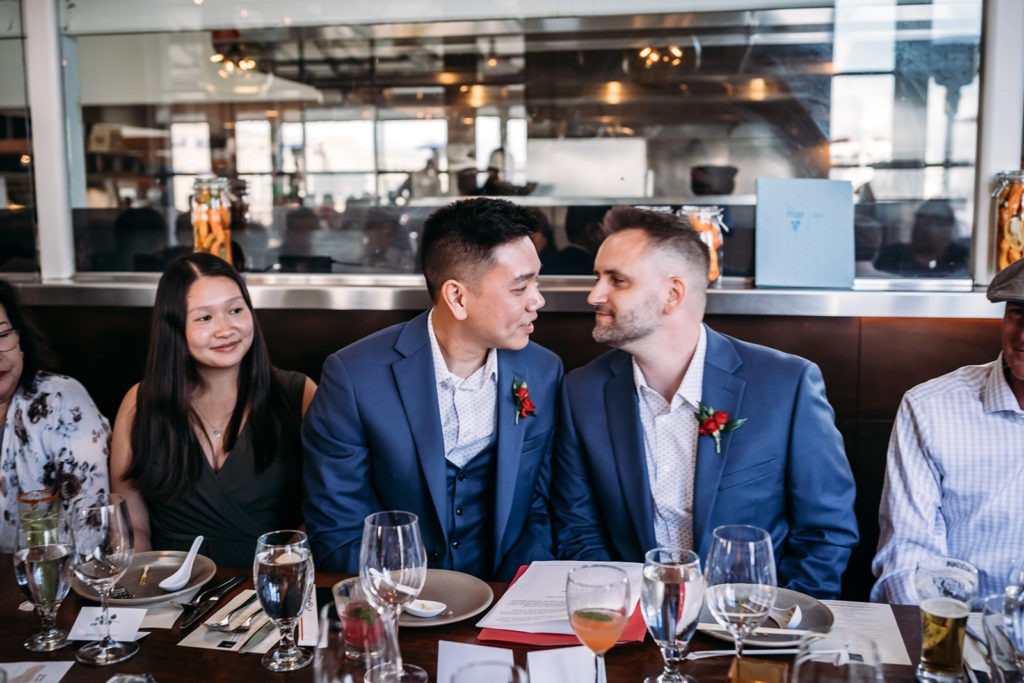 An inclusive and intimate wedding and elopement photographer, Xilo Photography, Kati Douglas who is based in Oakland, California in the bay area, took this picture of a same sex gay lgbtqia+ couple on their wedding elopement day as the two grooms celebrate and lean in for a kiss at the restaurant with their family and loved ones. This took place just after leaving their intimate wedding venue and was an unconventional and unique reception option.  Ever wondered what is an intimate wedding or what would be considered a small wedding? This is a great example of both! 