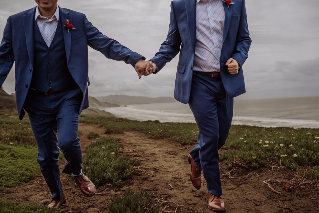 An intentional wedding and elopement photographer, Xilo Photography, Kati Douglas who is based in Oakland, California in the bay area, took this picture of a same sex gay couple on their wedding day/elopement day as the two grooms hold hands while running together in their tuxes at Fort Funston, CA which was their beach wedding venue. 