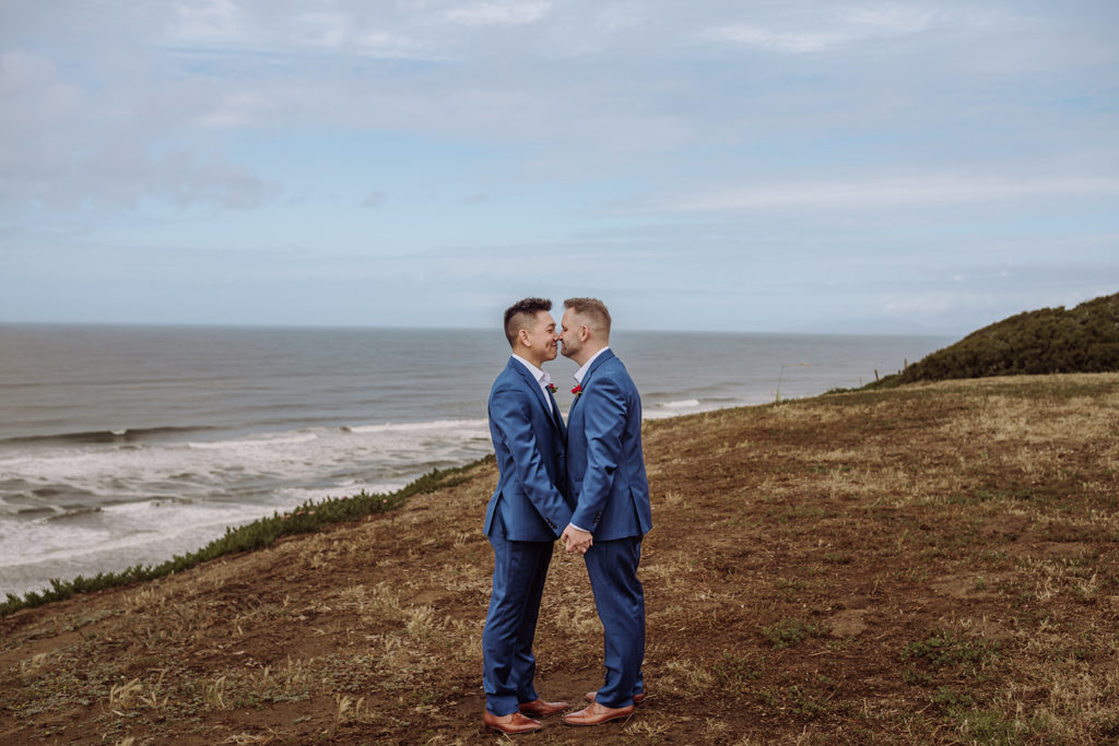 An inclusive intimate wedding and elopement photographer, Xilo Photography, Kati Douglas who is based in Oakland, California in the bay area, took this picture of a same sex gay couple on their wedding day/elopement day as the two grooms lean in and touch noses near the beach with waves crashing in the background at Fort Funston, CA which was their intimate wedding venue. Ever wondered what is an intimate wedding or what would be considered a small wedding? This is a great example of both! 