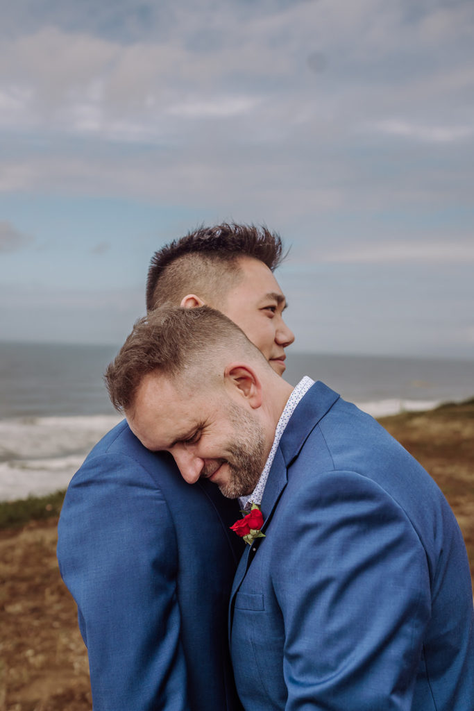 An inclusive wedding and elopement photographer, Xilo Photography, Kati Douglas who is based in Oakland, California in the bay area, took this picture of a same sex gay couple on their wedding day/elopement day as the two grooms lean in near the beach with waves crashing in the background at Fort Funston, CA which was their outdoor beach wedding venue. 