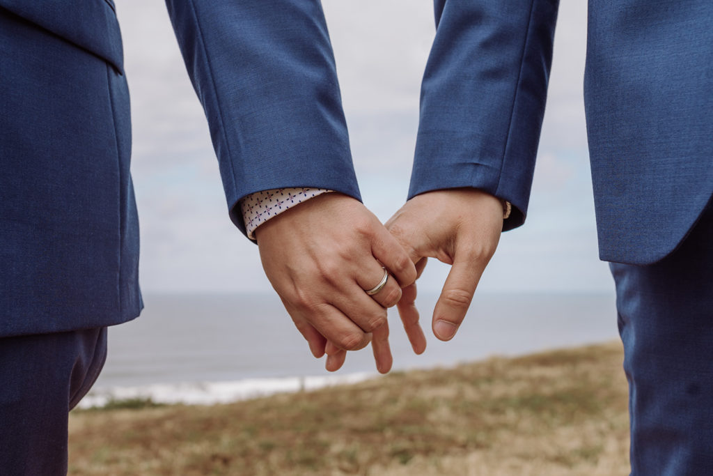 An inclusive wedding and elopement photographer, Xilo Photography, Kati Douglas who is based in Oakland, California in the bay area, took this picture of a same sex gay couple on their wedding day/elopement day as the two grooms hold hands while walking at Fort Funston, CA which was their outdoor wedding venue