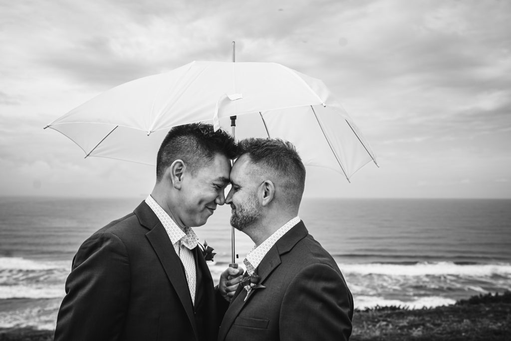 An inclusive intimate wedding and elopement photographer, Xilo Photography, Kati Douglas who is based in Oakland, California in the bay area, took this picture of a same sex gay couple on their wedding day/elopement day as the two grooms lean in and touch noses under an umbrella near the beach with waves crashing in the background at Fort Funston, CA which was their intimate wedding venue. Ever wondered what is an intimate wedding or what would be considered a small wedding? This is a great example of both! 
