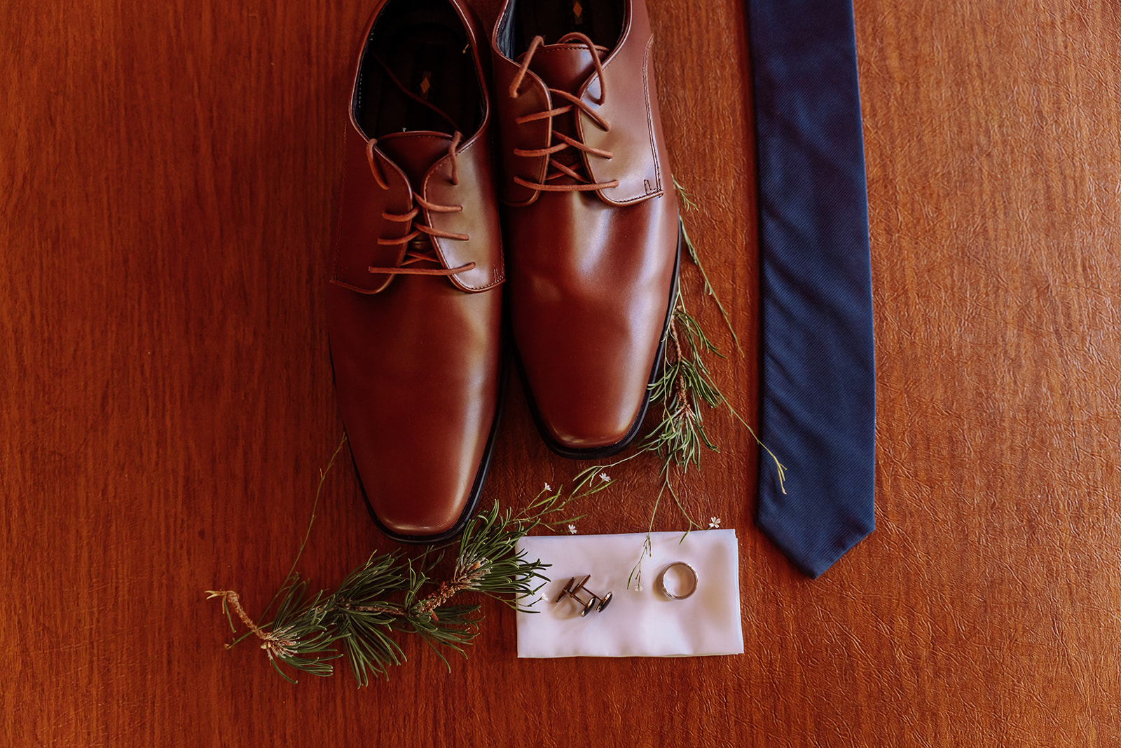Pictured here is a flat-lay of wedding keepsake details of the the groom's brown leather dress shoes, his navy blue tie, his ring and cuff links and hanker-chief on a gorgeous wood table. This was taken in Lake Tahoe, California during a mountain, forest wedding and outdoor forest beach ceremony. Photographed by Bay Area photographer, Kati Douglas with Xilo Photography based in Oakland, CA. 