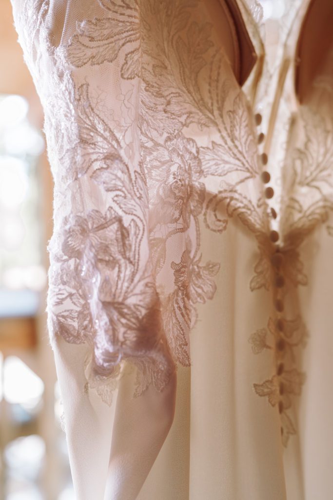 Gorgeous pristine A-Line Lace detailed wedding gown hung and focused in on the intricate beading, buttons, lace and detailing of this gown. This was taken in Lake Tahoe, California during a mountain, forest wedding and beach wedding that was actually a fairly intimate wedding venue, adorable romantic and natural wood decor, and outdoor forest beach ceremony. Photographed by Bay Area photographer, Kati Douglas with Xilo Photography based in Oakland, CA. 