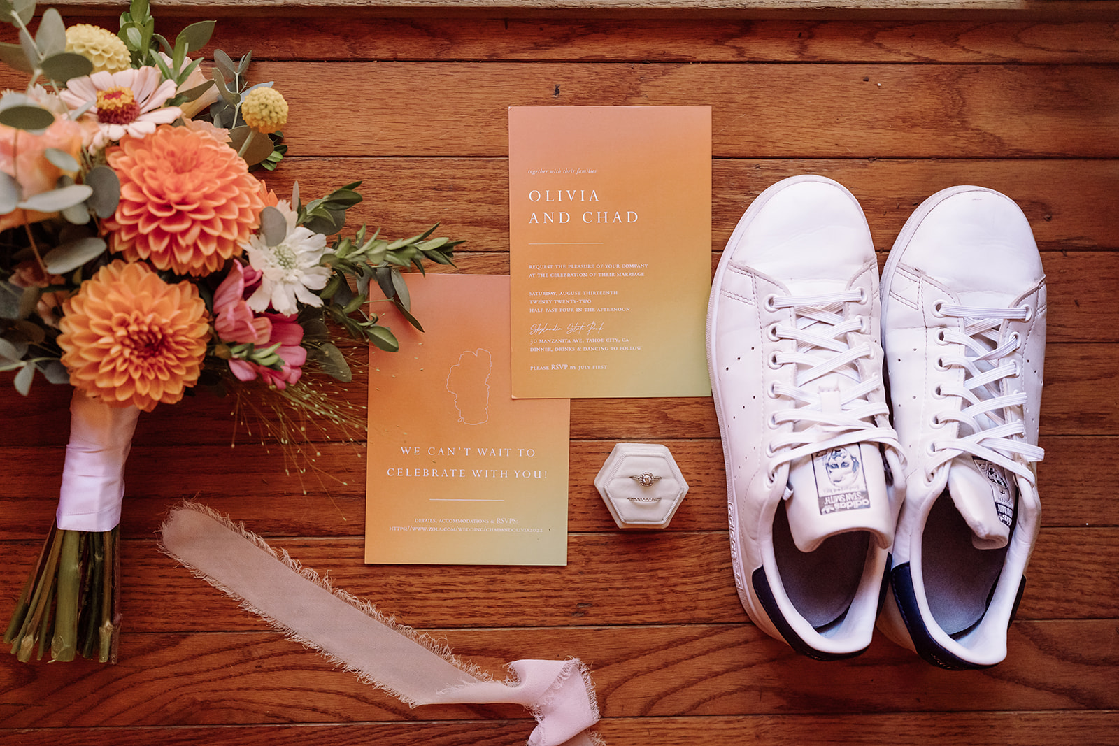 Pictured here is a flat-lay of wedding keepsake details of the bridal bouquet, white bridal sneakers, the bridal wedding ring, and sunset colored invitations on a gorgeous wood table. This was taken in Lake Tahoe, California during a mountain, forest wedding and beach wedding that was at an outdoor forest beach ceremony. Photographed by Bay Area photographer, Kati Douglas with Xilo Photography based in Oakland, CA. 