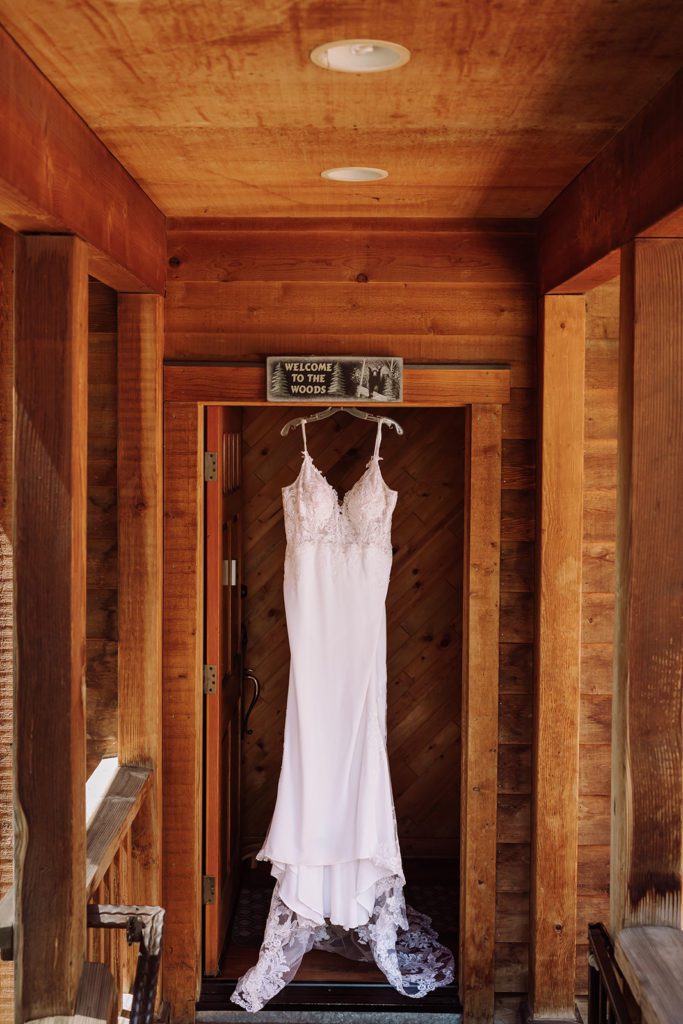 Gorgeous pristine A-Line lace detailed wedding gown hung and your eye is drawn to the intricate beading, buttons, lace and detailing of this gown. This was taken in Lake Tahoe, California during a mountain, forest wedding and beach wedding that was actually a fairly intimate wedding venue, adorable romantic and natural wood decor, and outdoor forest beach ceremony. Photographed by Bay Area photographer, Kati Douglas with Xilo Photography based in Oakland, CA. 