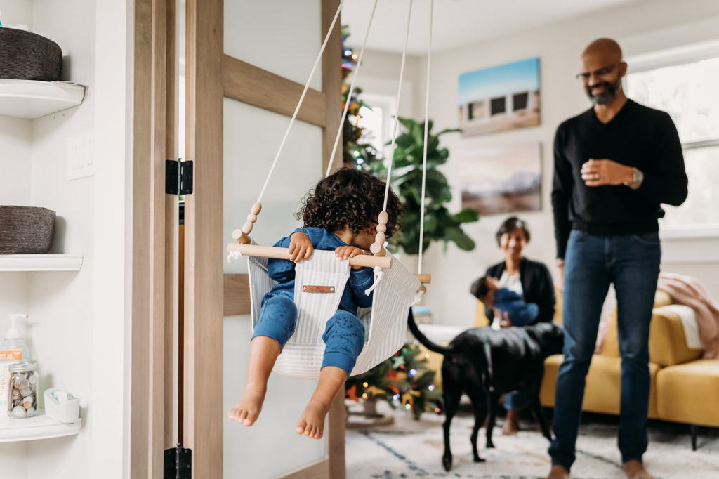 This sweet newborn documentary session at home in Oakland CA included mother, father, and their two children during this home session by Xilo Photography. The older toddler is in an indoor doorway swing and having a blast being pushed by their father and swinging.